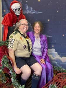 A woman in a Discworld Scout Uniform and another woman in a blue suit sit in the Hogfather's sleigh, with Death (dressed as the Hogfather) behind them