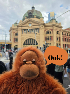 The Librarian (an orangutan toy) in front of Melbourne's Flinders Street Station. He has a word balloon saying "Ook!"