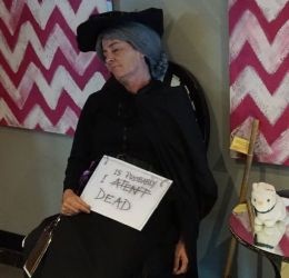 Granny Weatherwax sits in a chair, her eyes closed. She holds a sign saying "I Aten't Dead" although Aten't is crossed out with the words "Is Probably" above it. It has been a very long convention! (Nullus Anxietas 7, 2019)